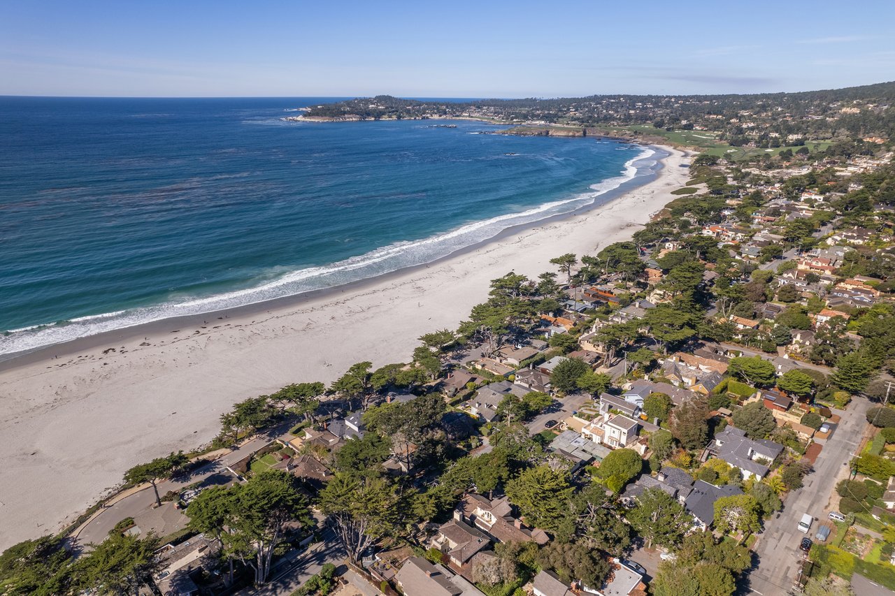 Aerial view of the crescent-shaped Carmel Beach with white sand, turquoise water, and coastal homes, looking toward the Pebble Beach golf course and Point Lobos in the distance