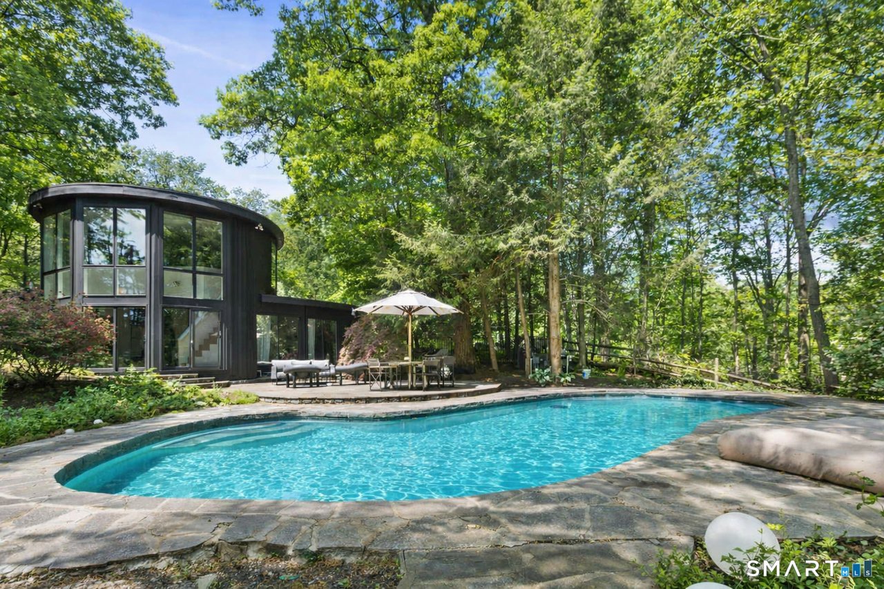 Swimming pool and stone patio beside a modern glass house in a wooded setting.