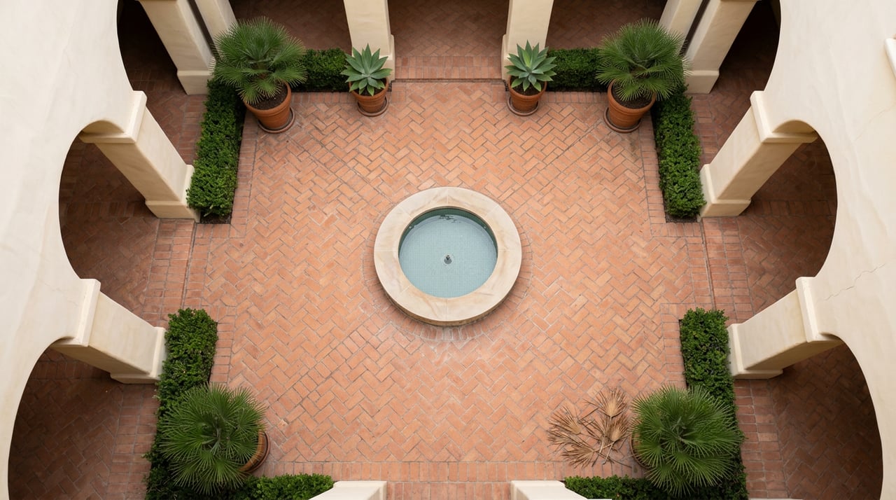 Enclosed courtyard with a round tiled fountain, herringbone terracotta paving, and potted palms beneath a plaster arcade.