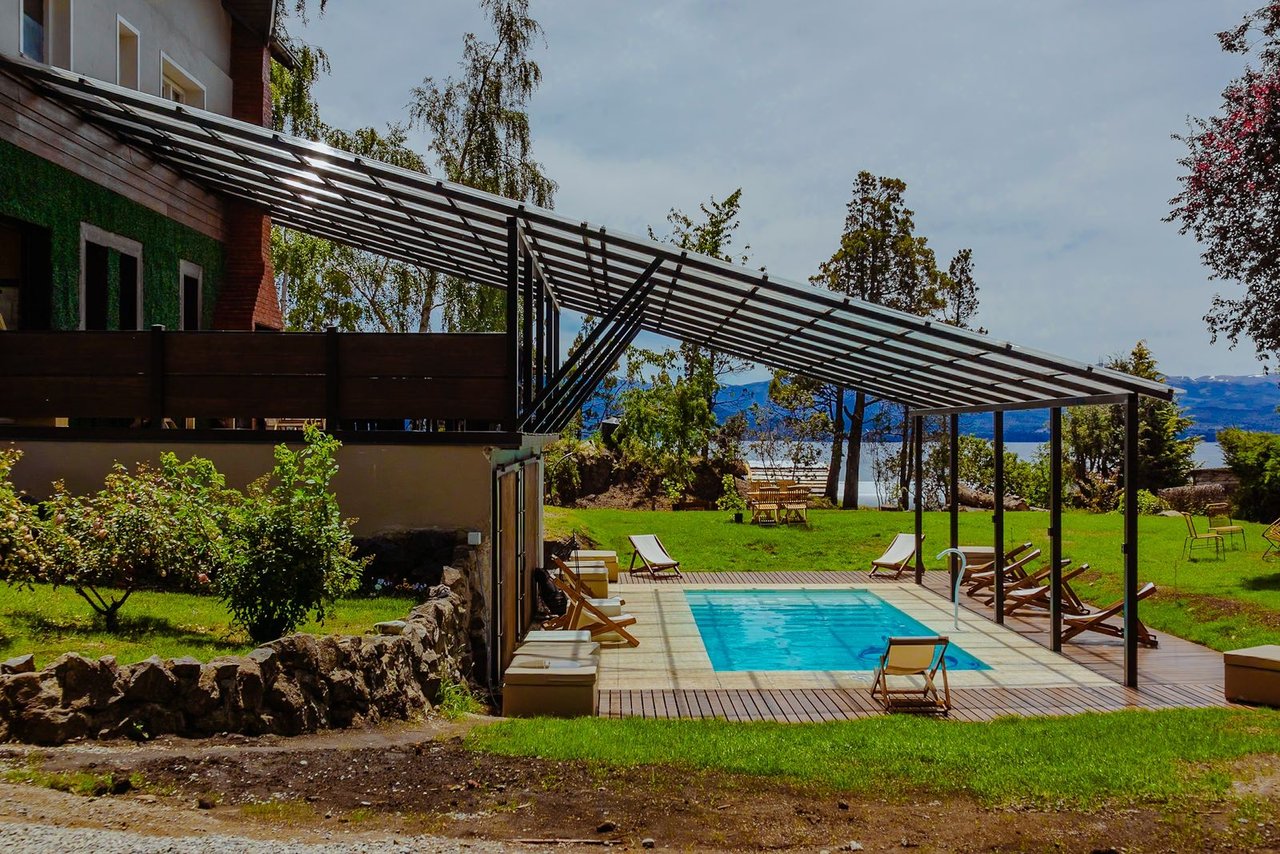Boutique hotel pool beneath a pergola, with a lake and mountains in the background.