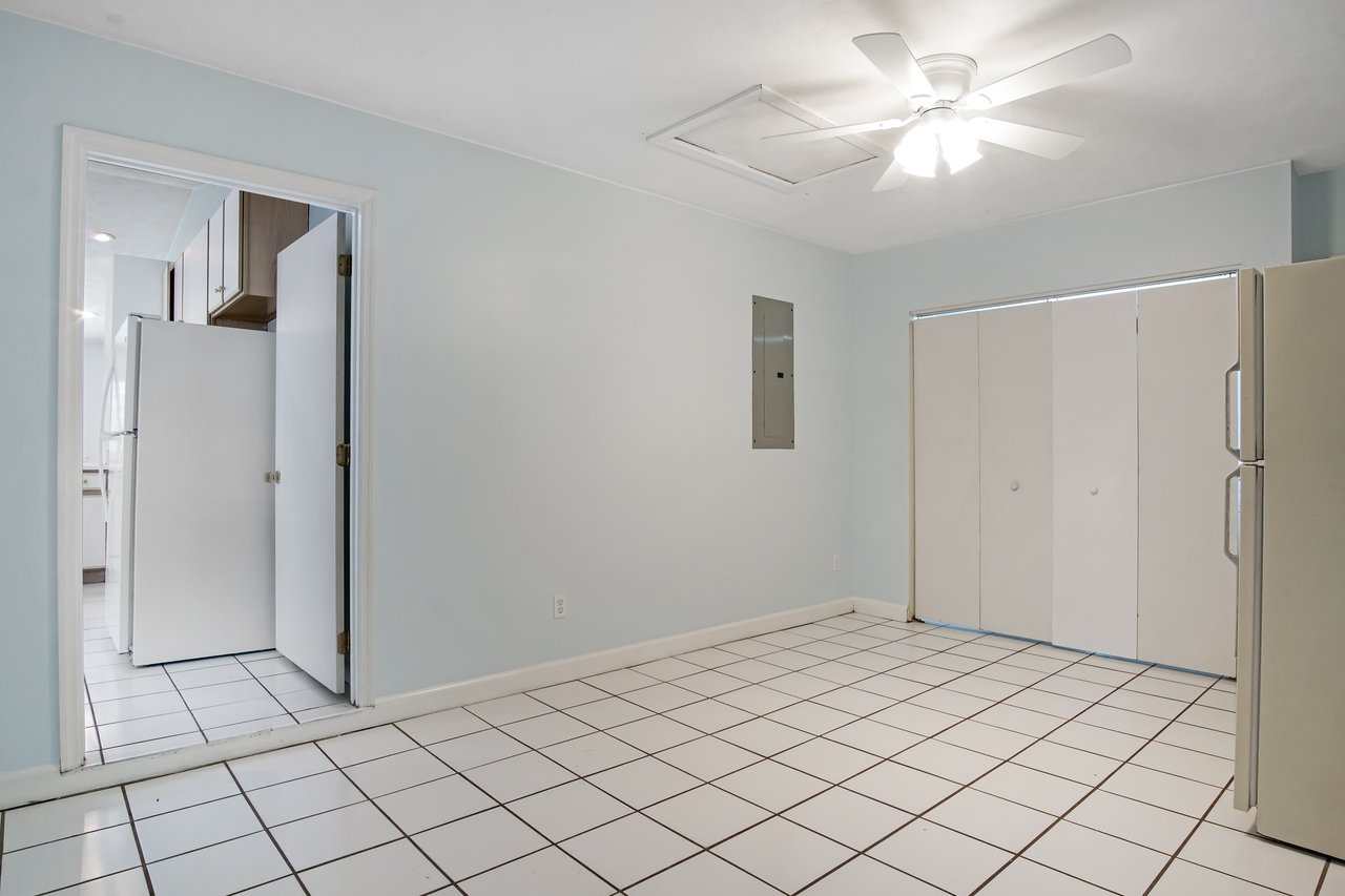 A tiled room with light blue walls, a ceiling fan, and a doorway leading to a kitchen with cabinets. White doors and a beige fridge suggest a clean, spacious feel.