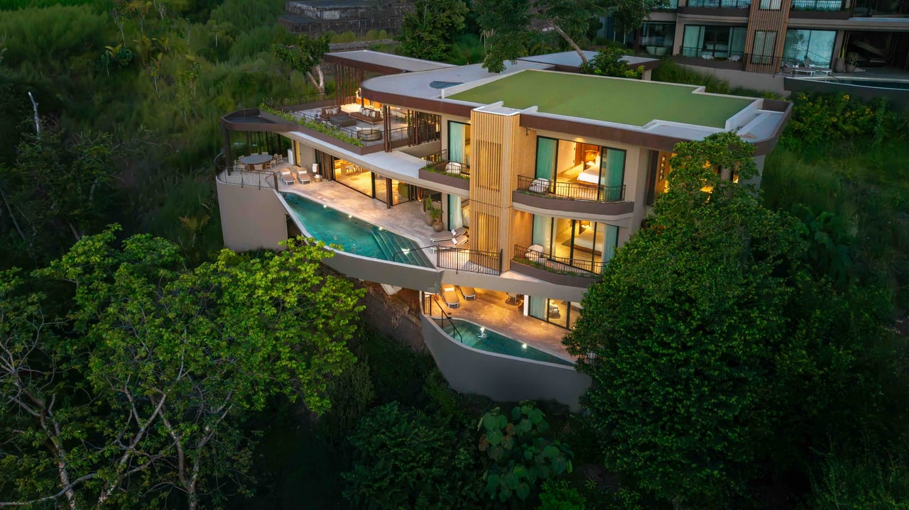 Modern multi-level estate home with curved terraces and a pool, surrounded by tropical greenery at dusk.