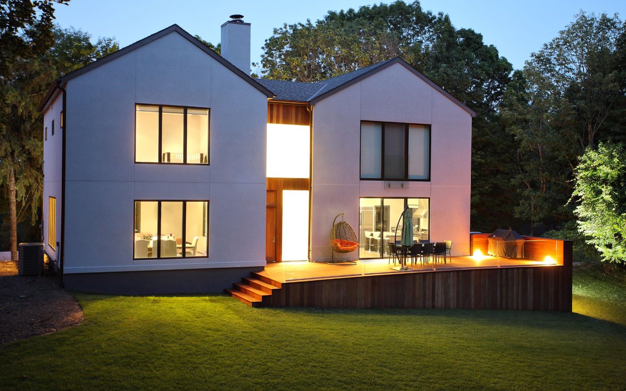 Modern two-story home glowing at dusk, with a raised wood deck, broad lawn, and mature trees.
