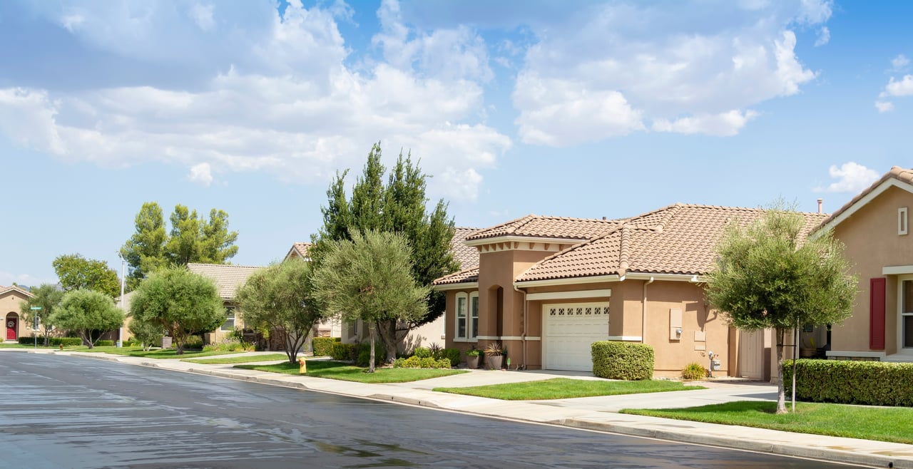 Residential street with single-story stucco homes, tiled roofs, and mature trees.