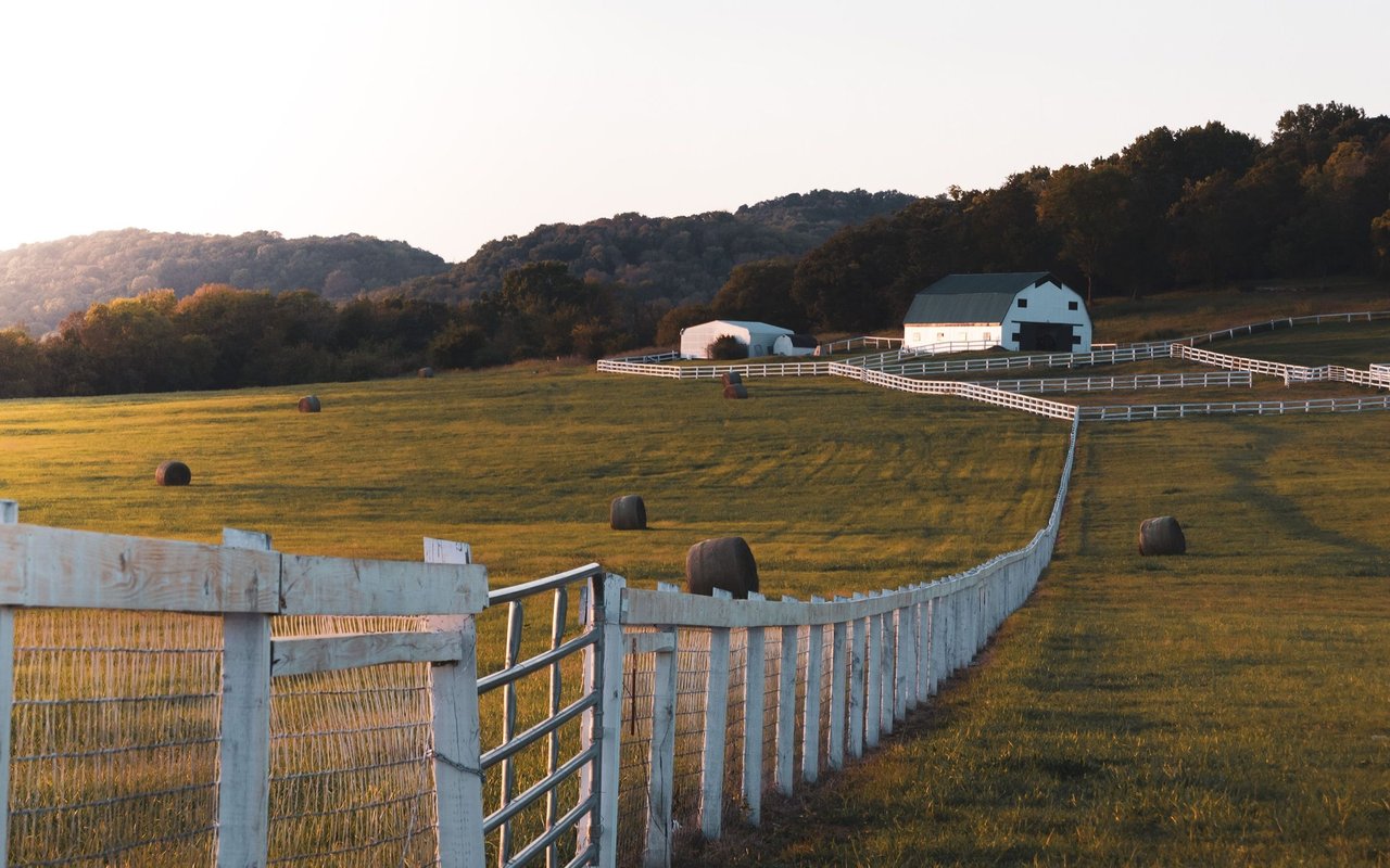 Fenced pasture with hay bales and a white barn beneath rolling, tree-covered hills.