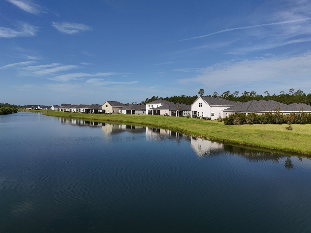 Homes line a grassy lakeshore, reflected in the water beneath a blue sky.