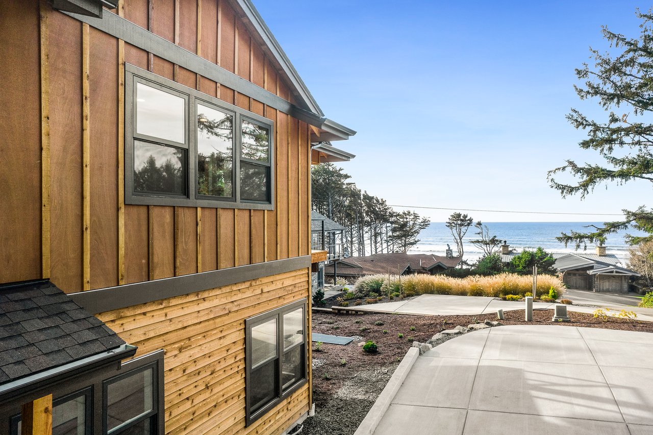 Wood-sided coastal home with a paved driveway and ocean visible beyond the neighboring homes.
