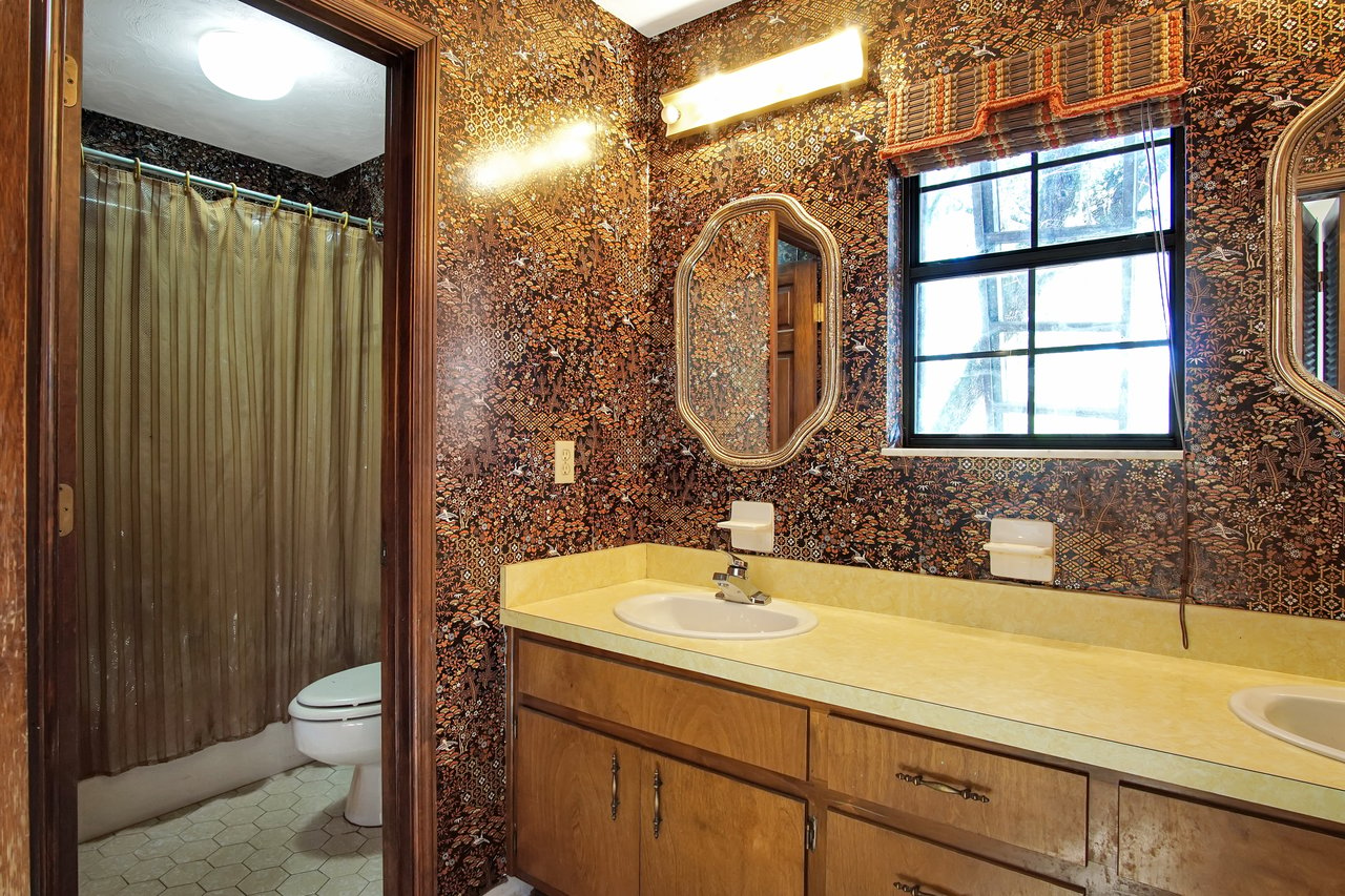 Vintage bathroom with bold, patterned wallpaper. A double sink vanity with yellow countertop, framed mirrors, and a small window. Warm, retro ambiance.