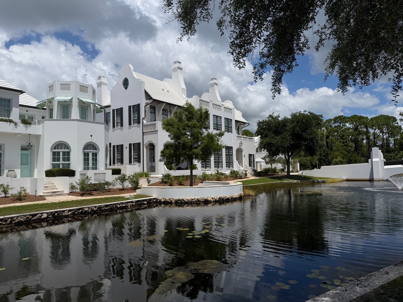 White luxury masonry home with dark shutters reflecting in a scenic pond with a fountain in Alys Beach.