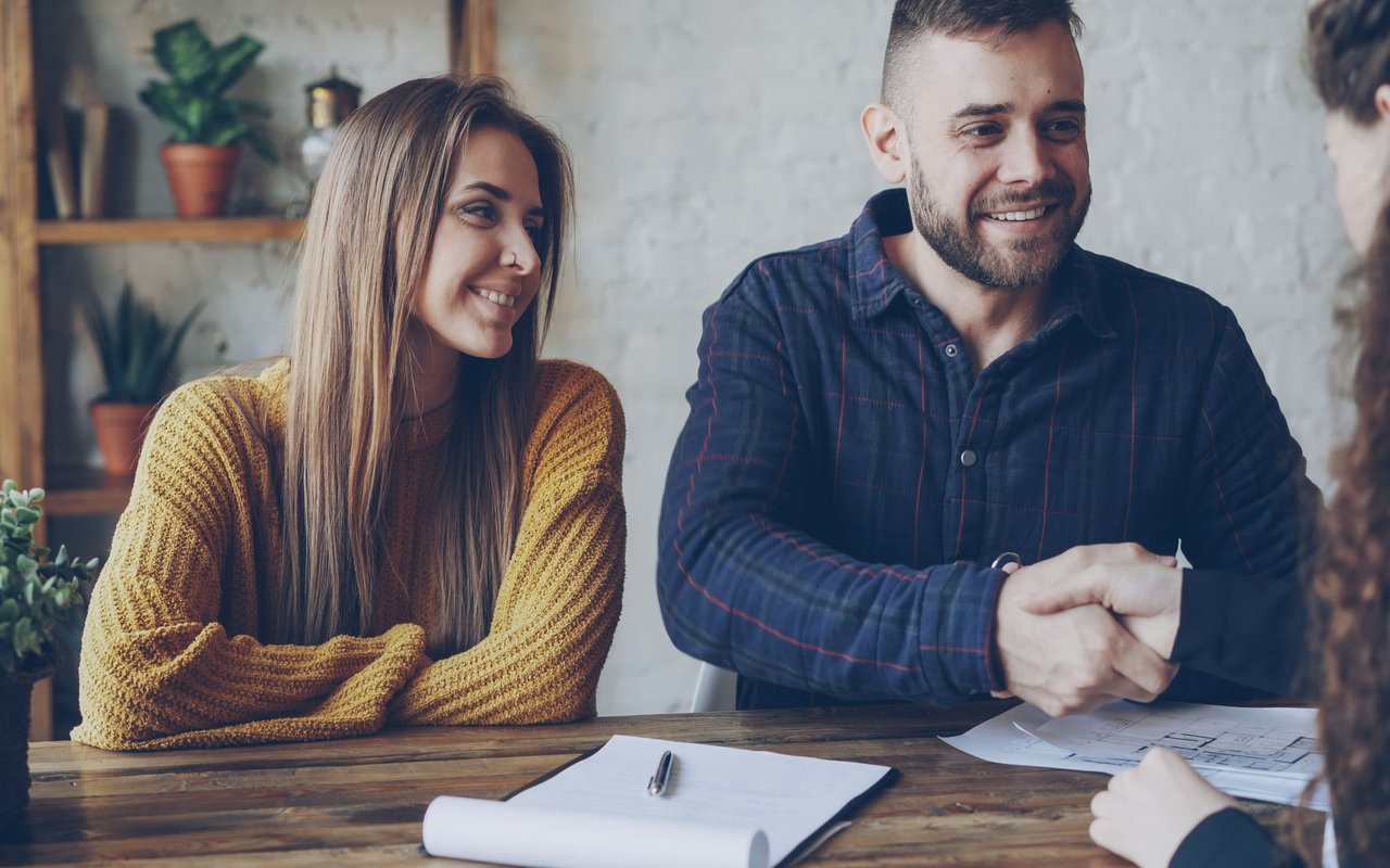 A smiling couple reviews paperwork with another person across a wooden table.