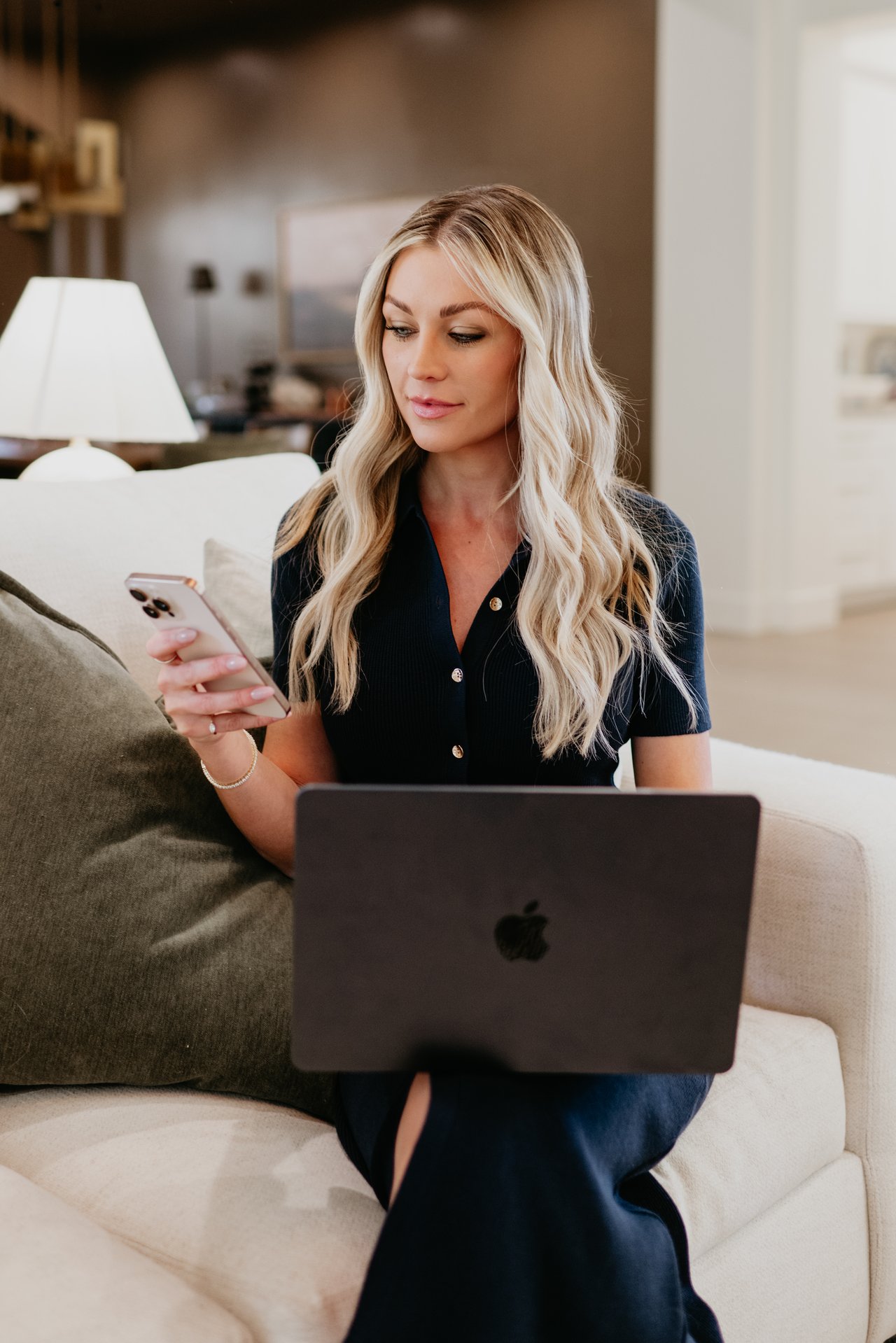 Woman on a sofa checking her phone beside an open laptop.