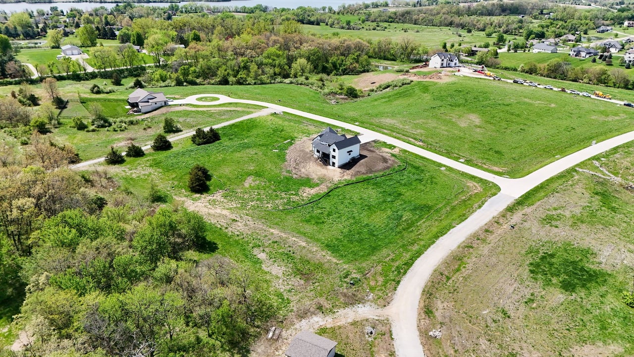 Aerial view of green development lots and curving roads, with a newly built home near the center.