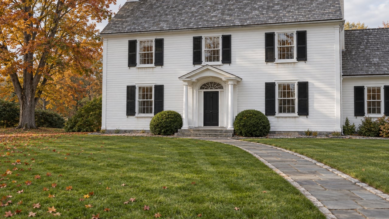 White clapboard home with black shutters, a deep lawn, stone entry path and amber autumn maple.