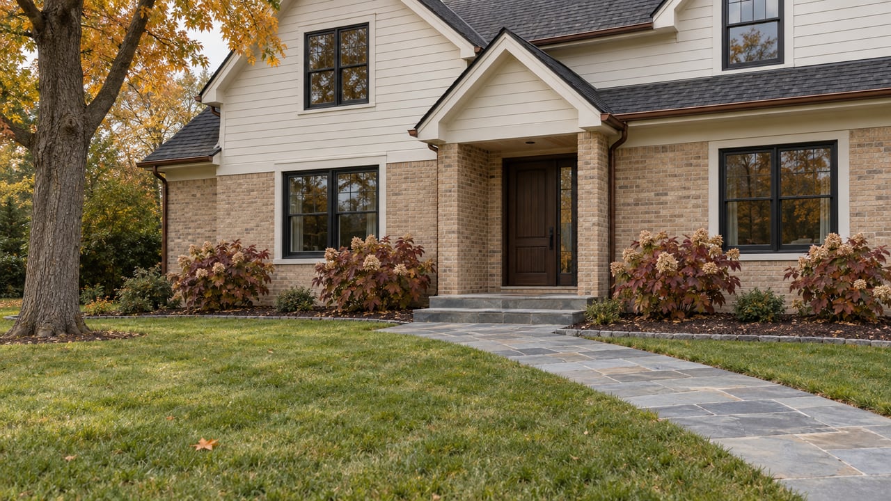 Updated gabled home with ivory siding, buff brick, black-trim windows, a bluestone front walk, and autumn maple foliage across a broad lawn.