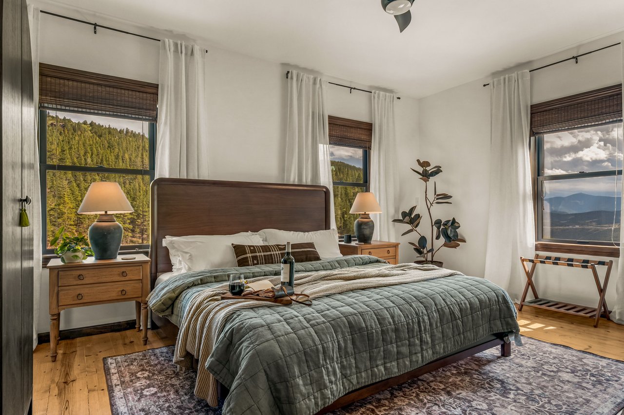Bedroom with a green quilted bed, wooden furnishings, and windows overlooking forested hills and mountains.
