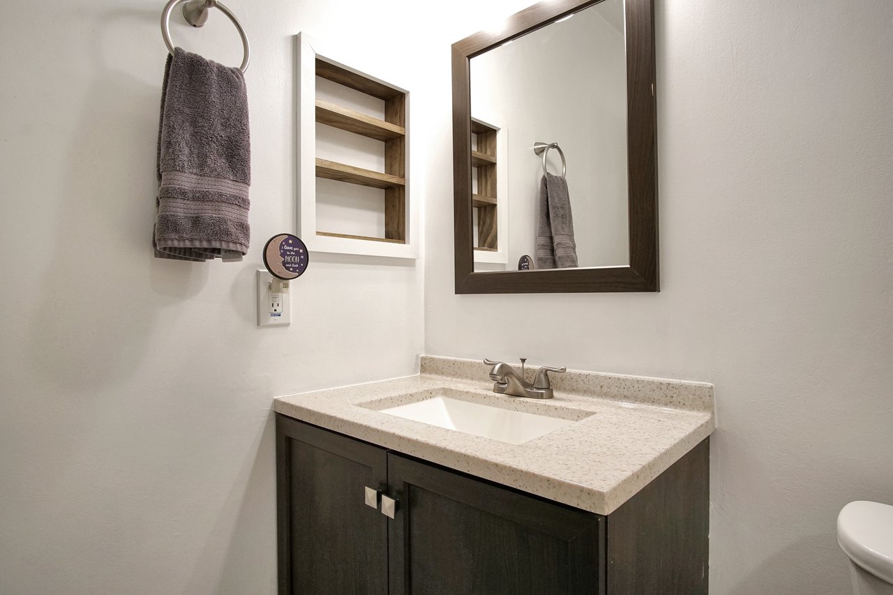 Modern bathroom with a dark wood vanity and beige countertop. A rectangular mirror is above the sink, flanked by a towel on a ring and open shelves.