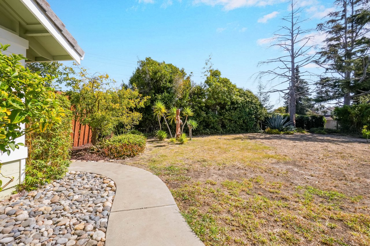 Backyard with a curved walkway, river-rock border, shrubs, and dry lawn.