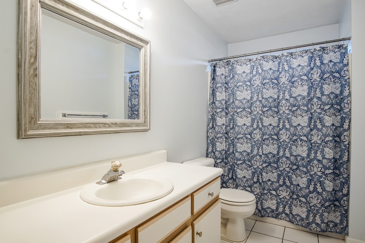 Bathroom with a large mirror, cream-colored countertop, and sink. A toilet sits beside a blue patterned shower curtain, creating a calm, clean ambiance.