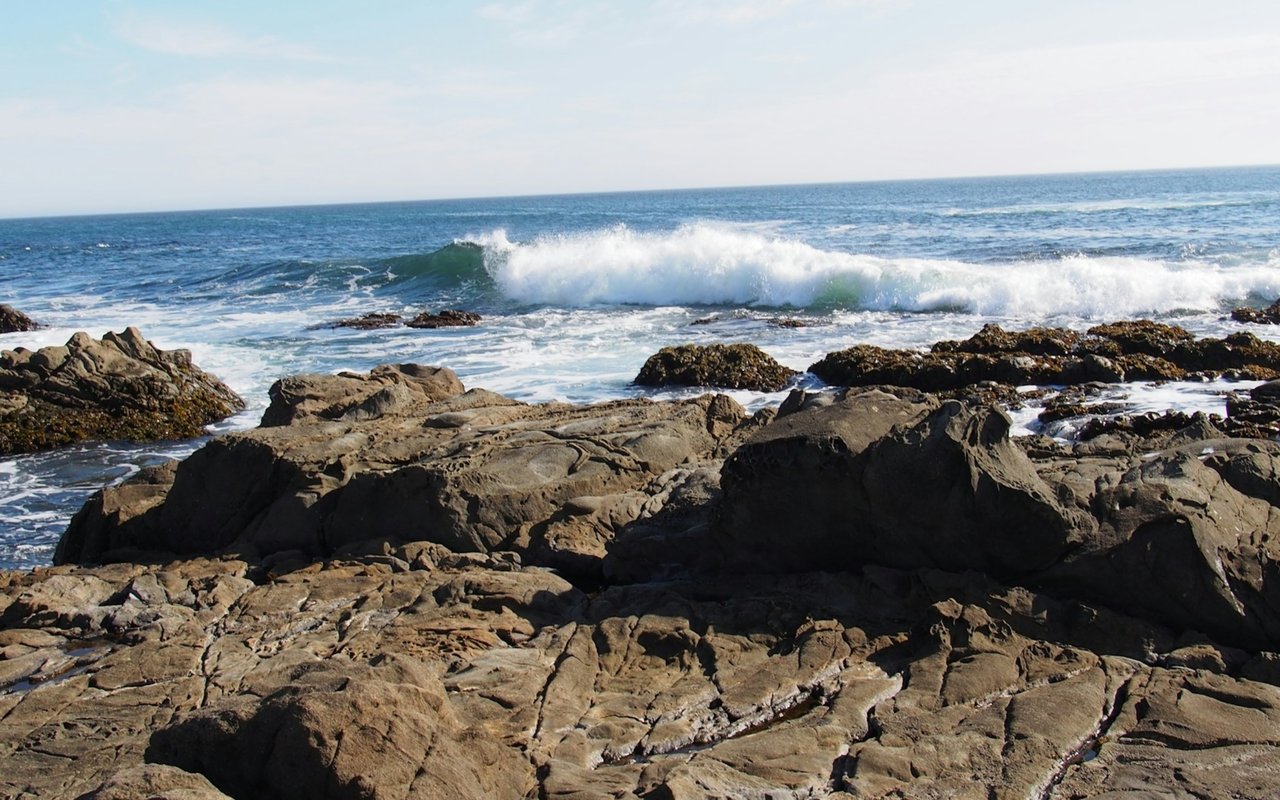 Breaking wave washes over a rocky shoreline beneath a pale blue sky.