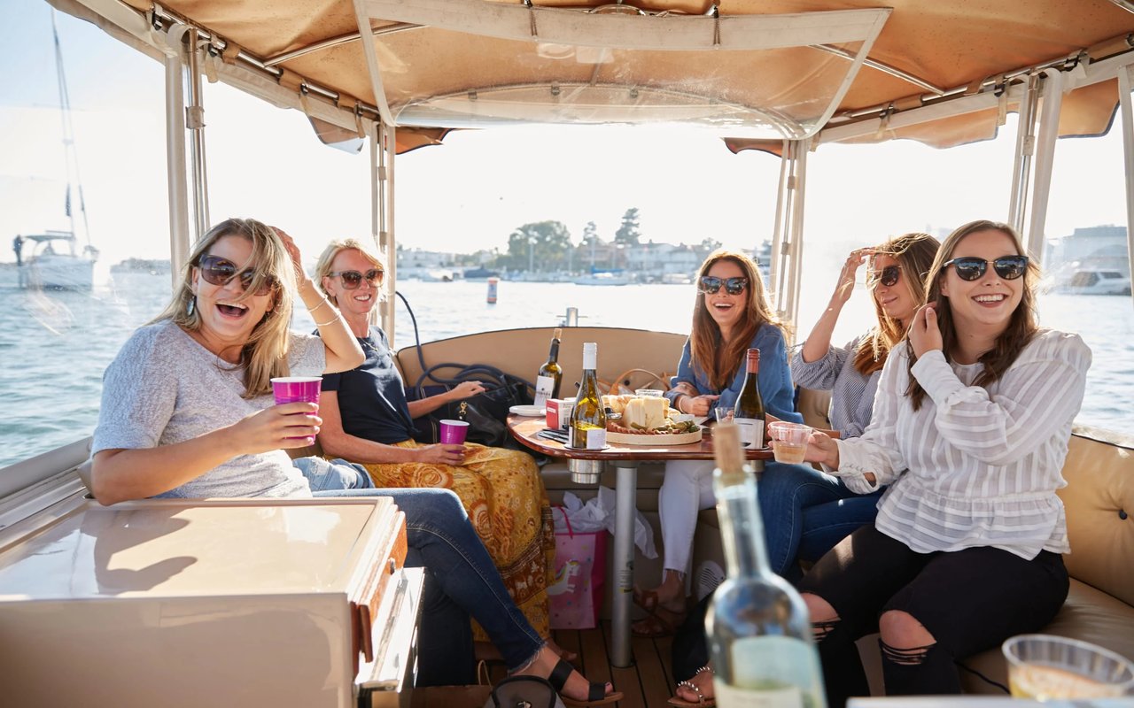A group of women laughing and smiling on a boat, enjoying drinks and snacks on a sunny day.