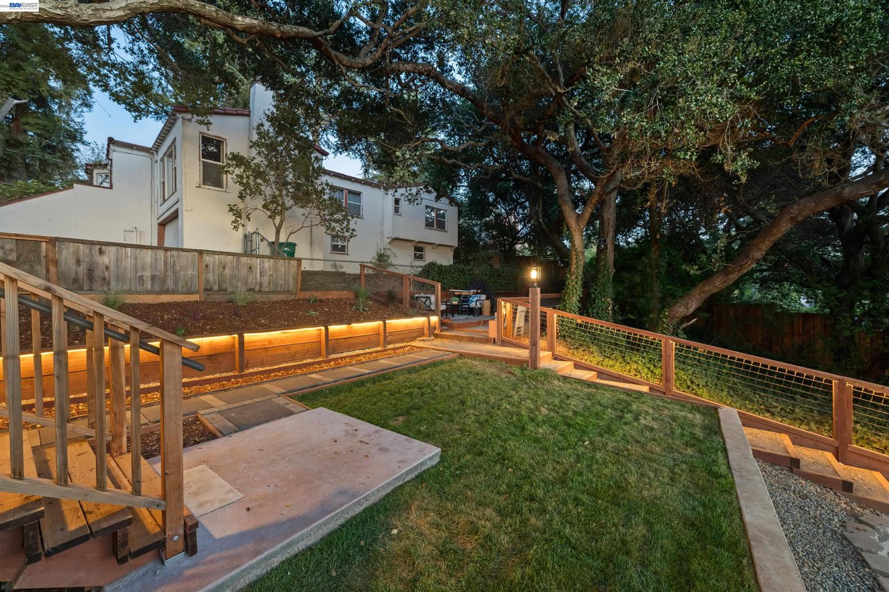 White multi-level home behind a landscaped backyard with timber railings, raised beds, and a large shade tree.