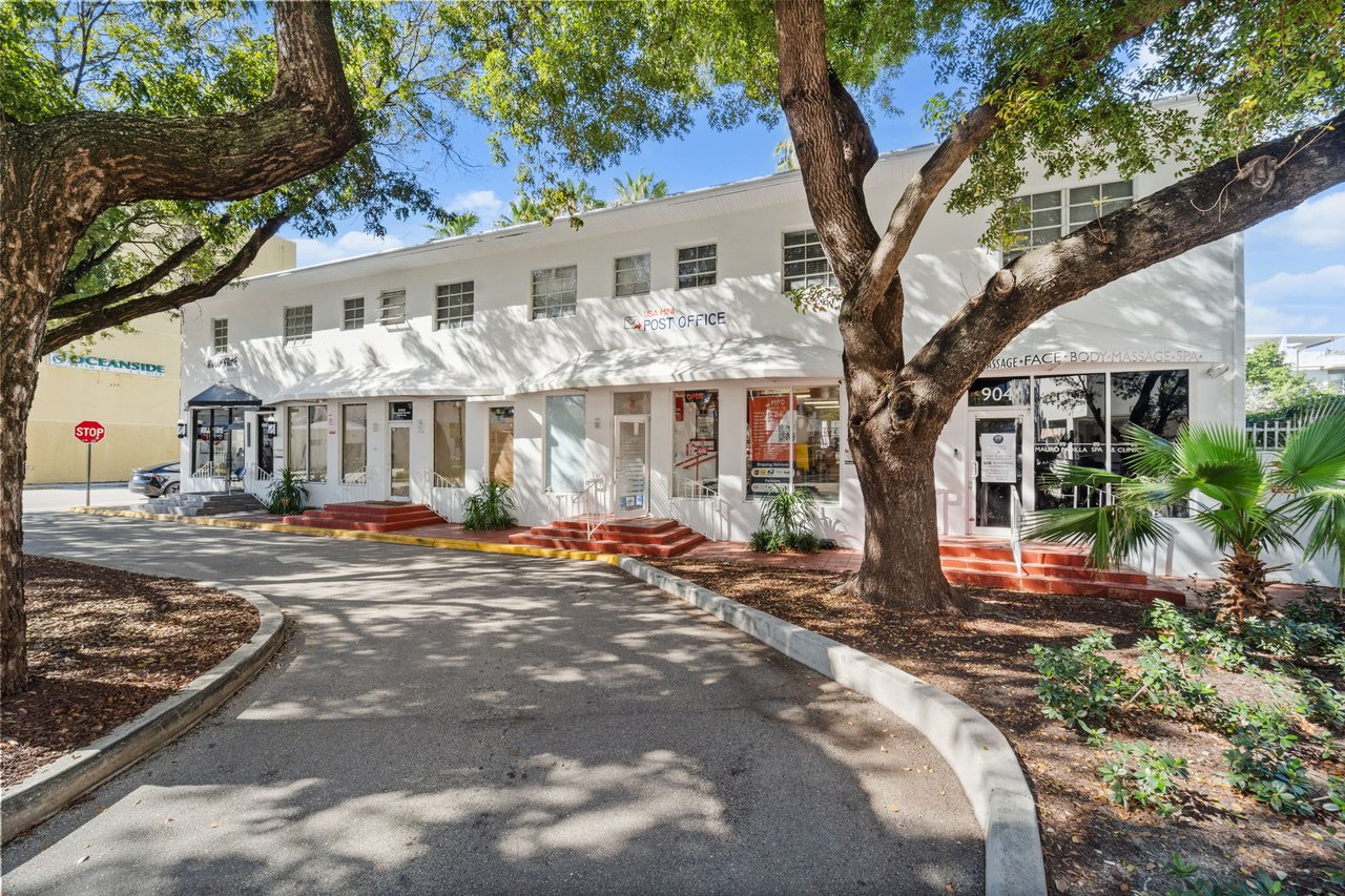 White commercial building with storefronts and a post office entrance, shaded by large trees.