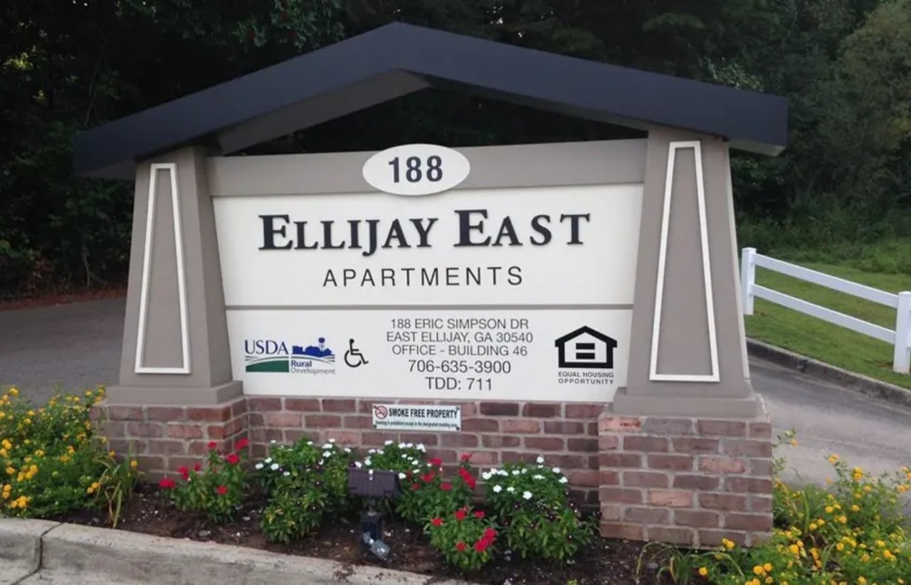 Ellijay East Apartments entrance sign on a brick base beside flower beds and a driveway.