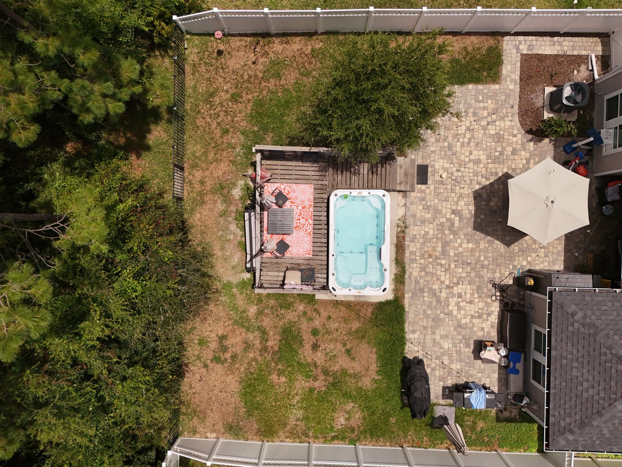 Fenced backyard with a paved patio, hot tub, wooden deck, umbrella, and mature trees.