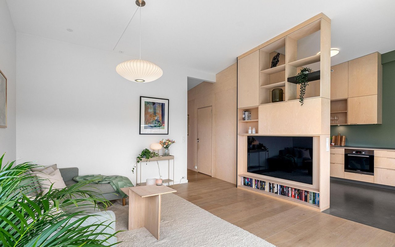 Open-plan living room with a gray sofa and pale wood cabinetry surrounding a built-in television.