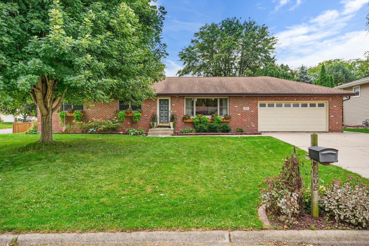 Front exterior of the red brick ranch at 1205 Wexford Drive in Waunakee’s Castle Crest neighborhood, featuring an attached two-car garage, mature trees, and landscaped front yard.