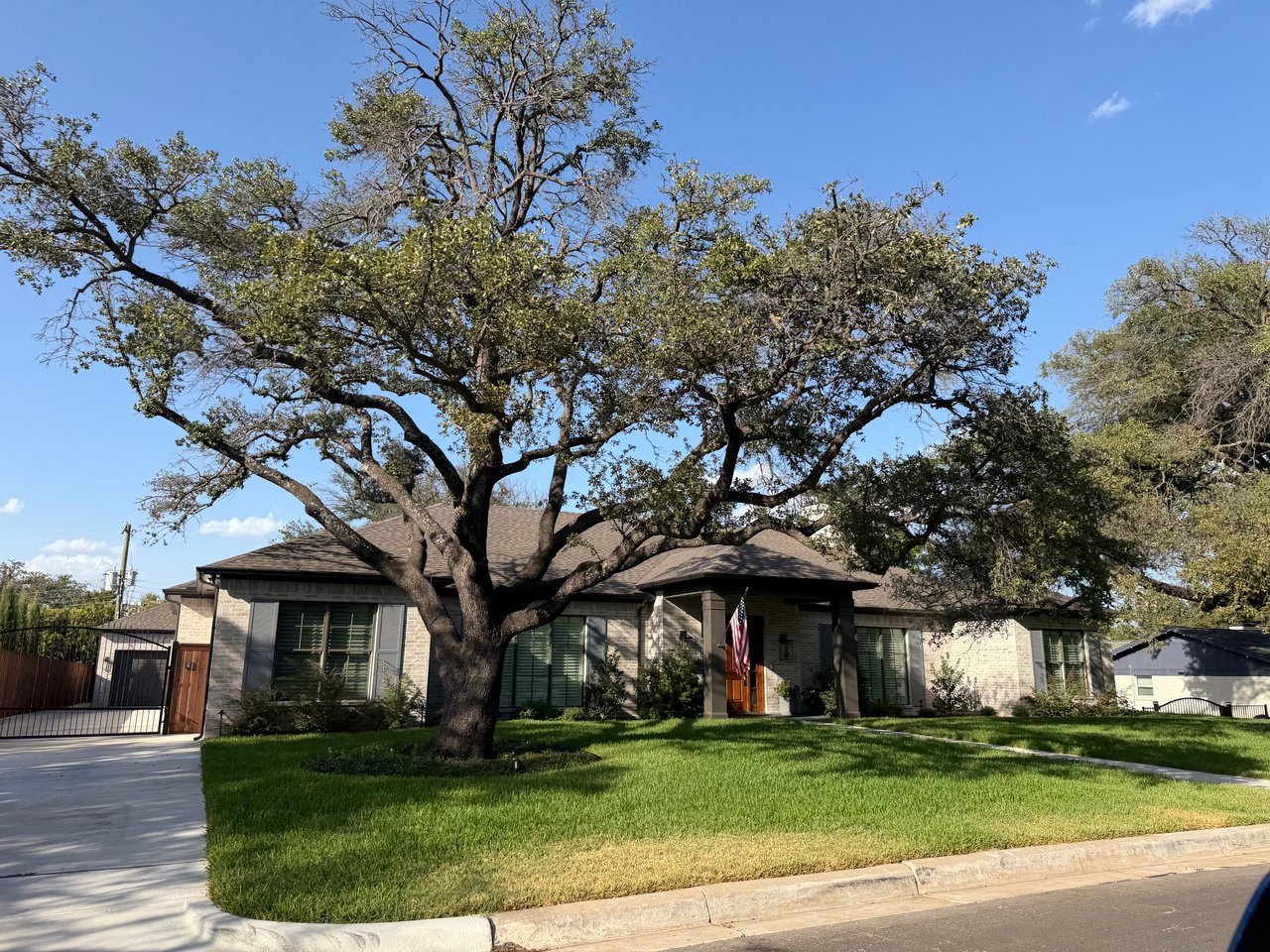 Single-story brick home with a large shade tree, manicured lawn, and American flag by the front entry.