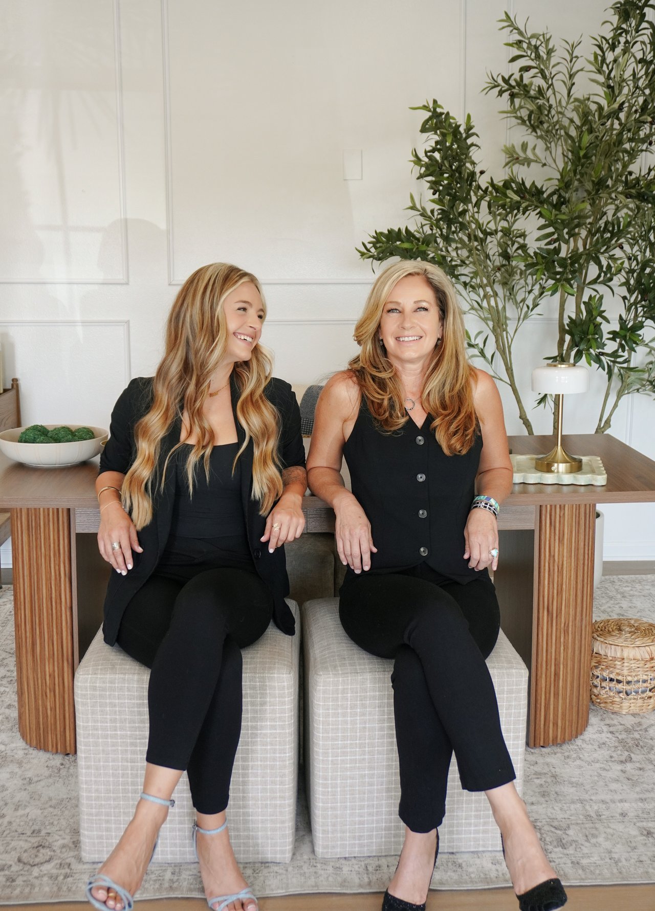 Two women in black seated side by side, smiling in a bright office interior.