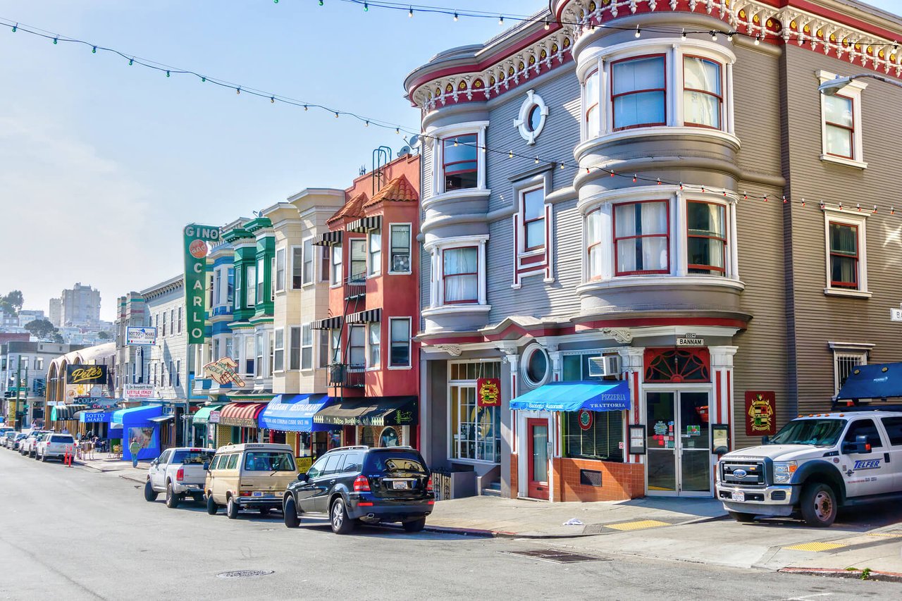 Street view of North Beach, San Francisco, featuring colorful historic buildings with bay windows, Italian restaurants, and local businesses. A mix of parked cars and pedestrians line the street under hanging string lights, creating a lively neighborhood atmosphere.