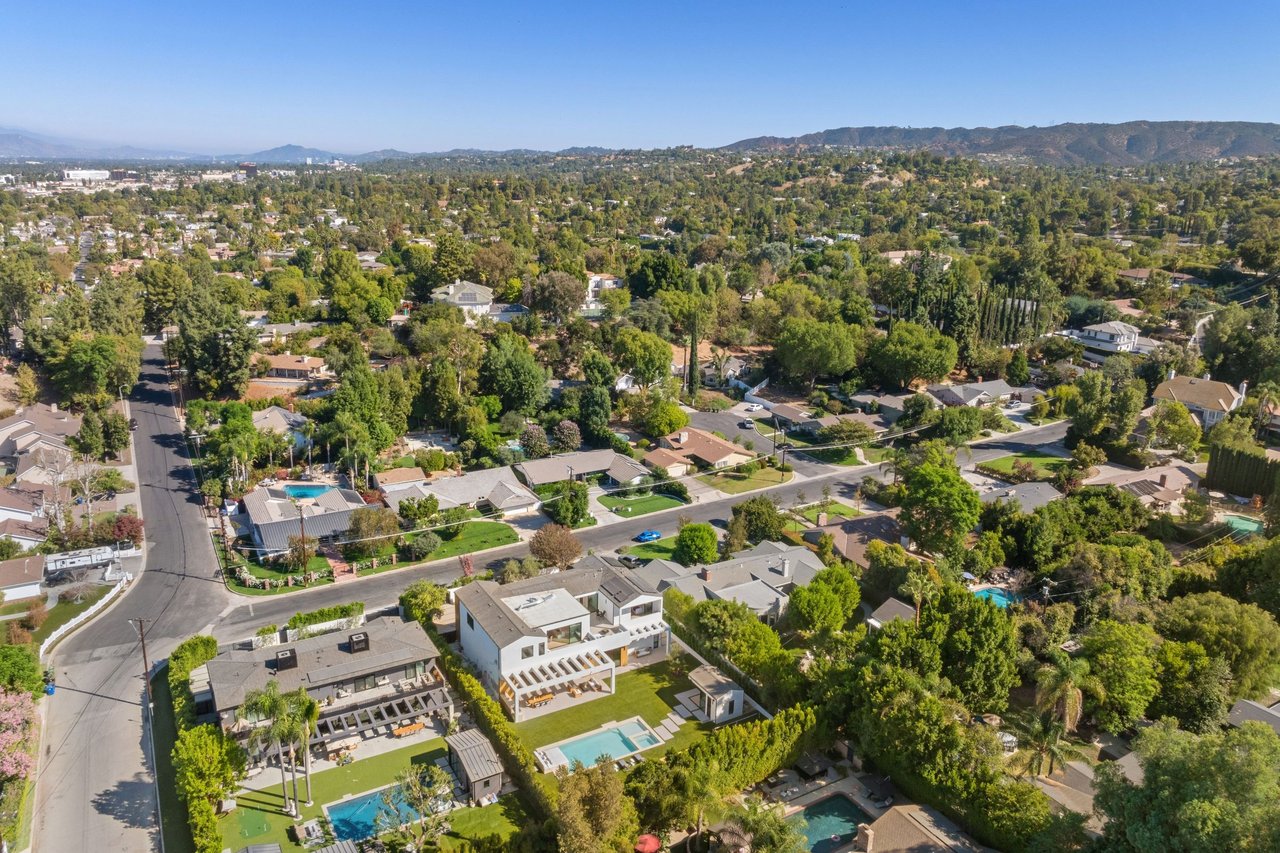 Aerial view of a leafy neighborhood with detached homes, swimming pools, and distant hills.