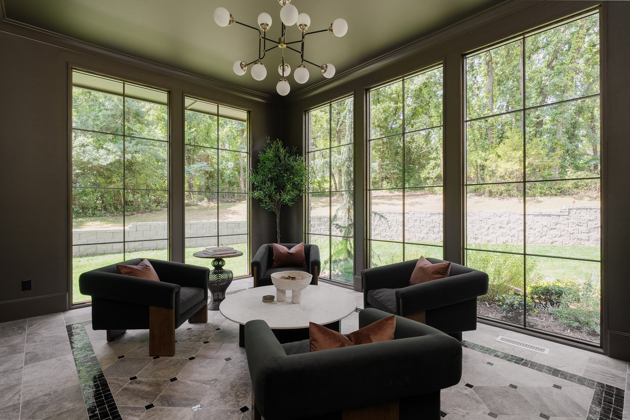 Furnished sitting room with tall grid windows overlooking trees and a stone retaining wall.