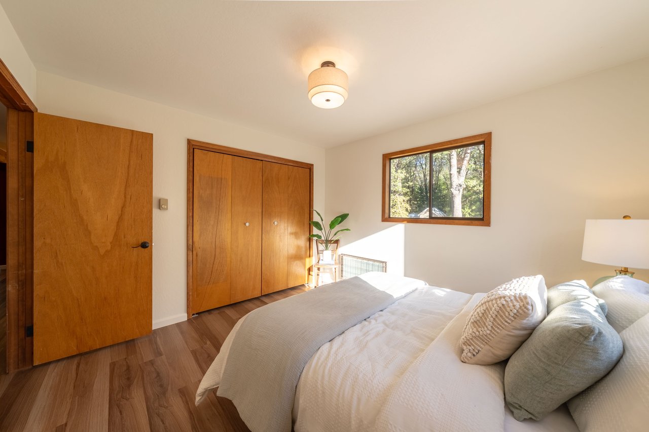 Bedroom with white bedding, wood closet doors, and a sunlit window overlooking trees.