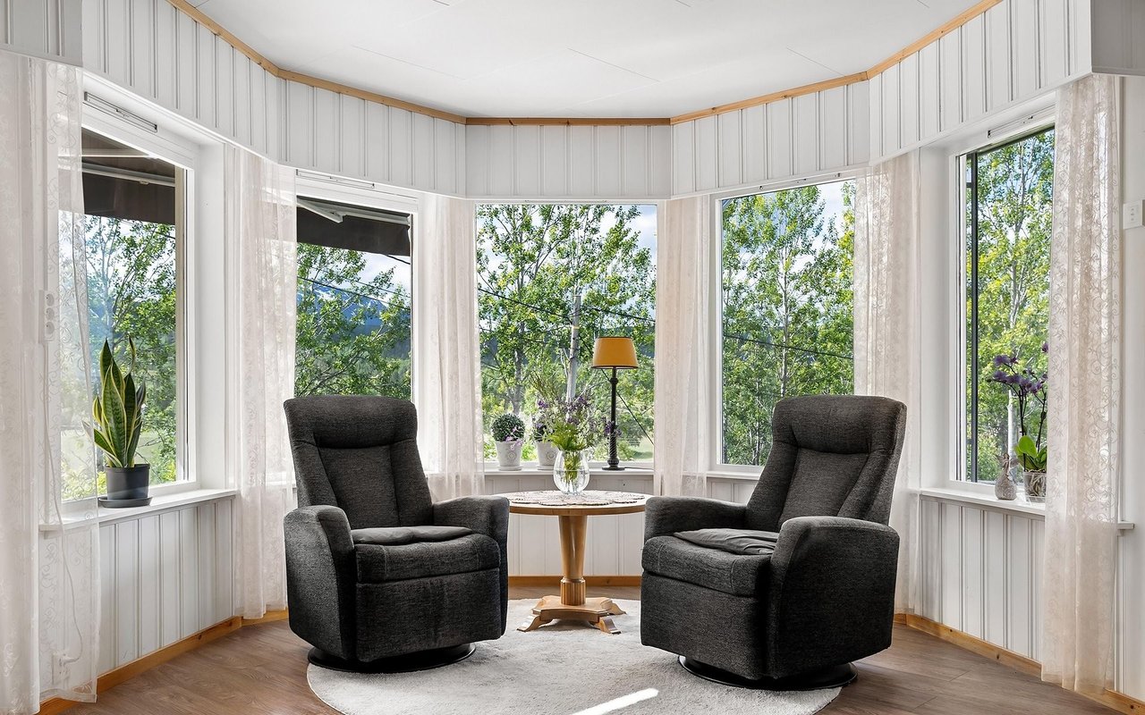 Sunroom with two dark recliners, a round table, and bay windows overlooking leafy trees.