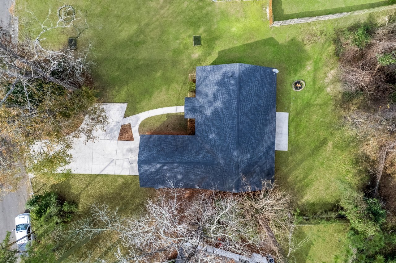 Aerial view of a single-story house with a dark roof, surrounded by a spacious green lawn, trees, and a driveway. A white car is parked on the street nearby.