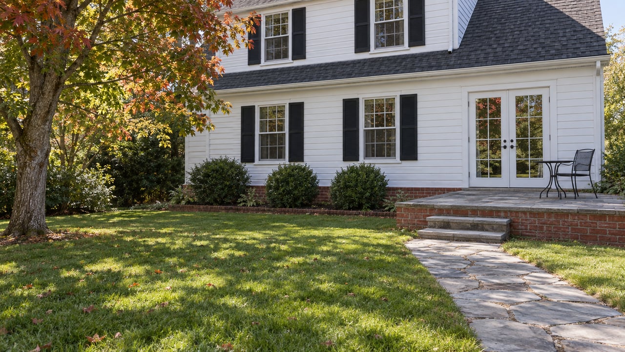 White clapboard two-story home overlooking a broad lawn, with paired black shutters and a raised brick-and-flagstone garden terrace.