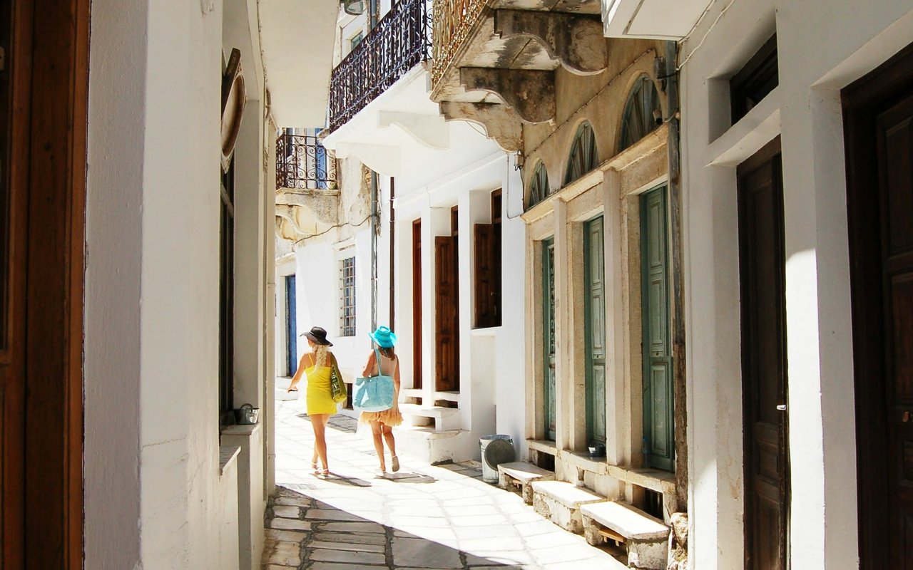 Two women walk through a sunlit alley lined with stone buildings and green shutters.