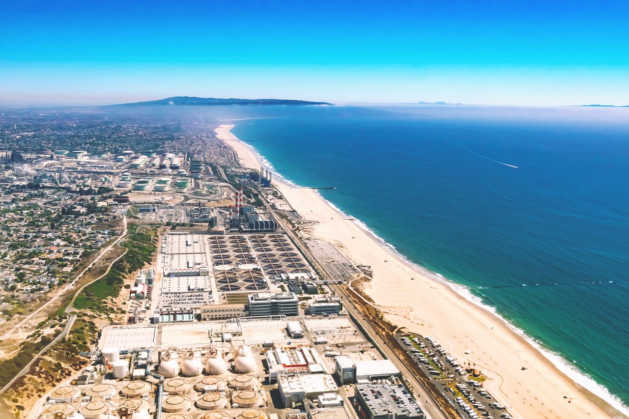 Aerial coastline with a broad sandy beach, blue ocean, and industrial facilities beside the shore.
