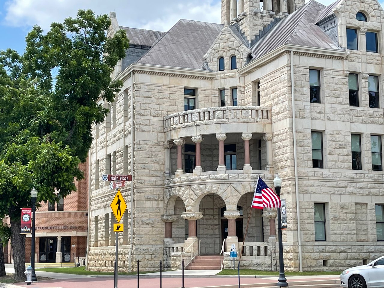 Image of the renovated Comal County Courthouse with the American flag flowing in the wind. There is a large pecan tree on the left and blue skies in the background.
