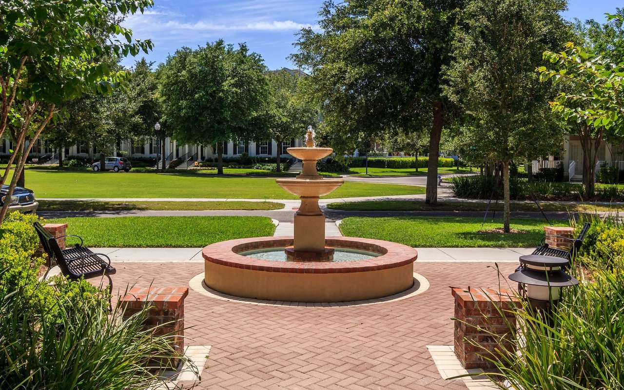 Neighborhood park fountain surrounded by brick paving, benches, green lawns, and mature trees.