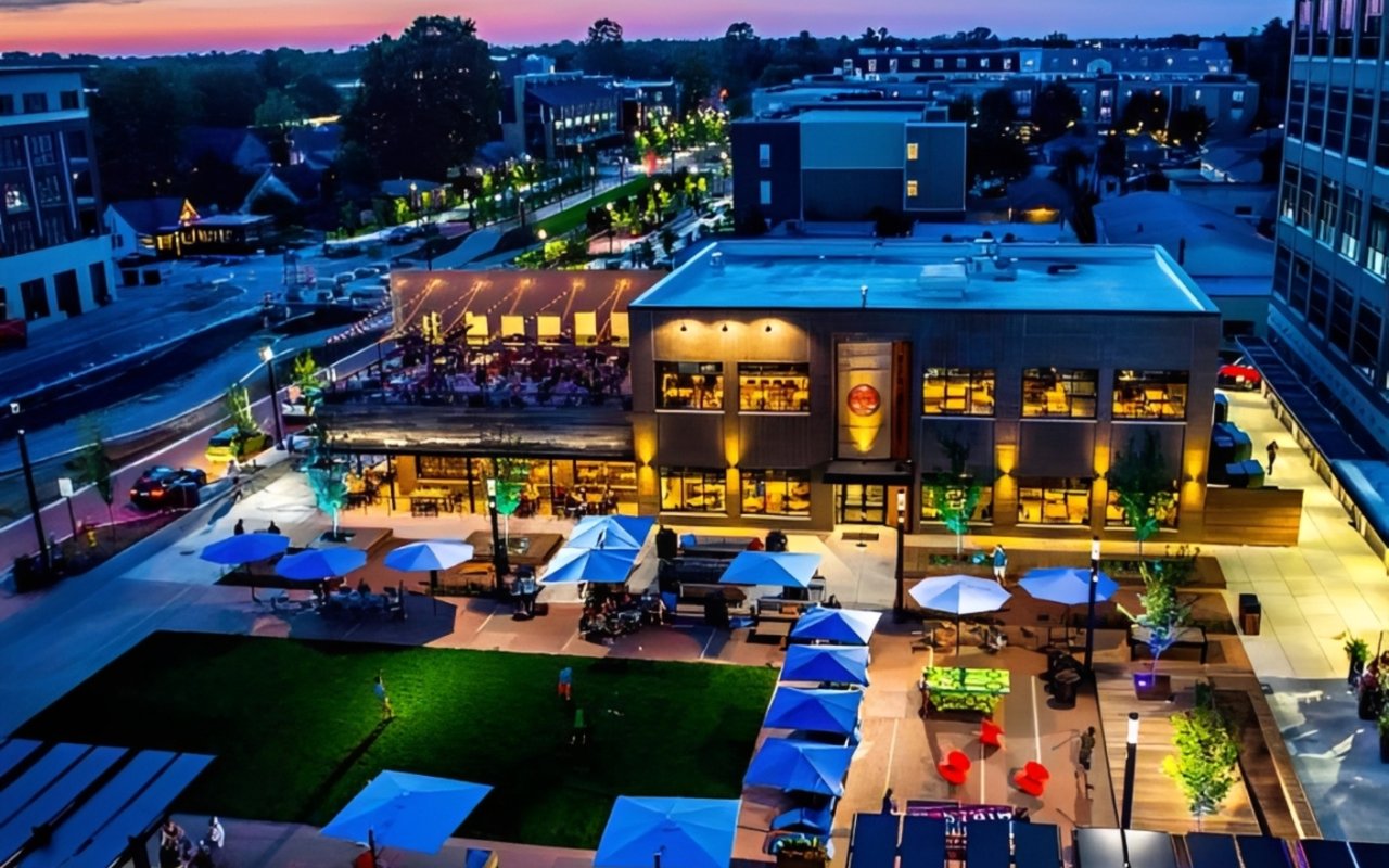 Downtown district at dusk with modern buildings, illuminated restaurants, and a plaza filled with blue umbrellas.