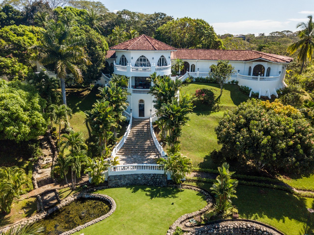 White villa with terracotta roofs, a grand staircase, tropical gardens, and stone-edged ponds.