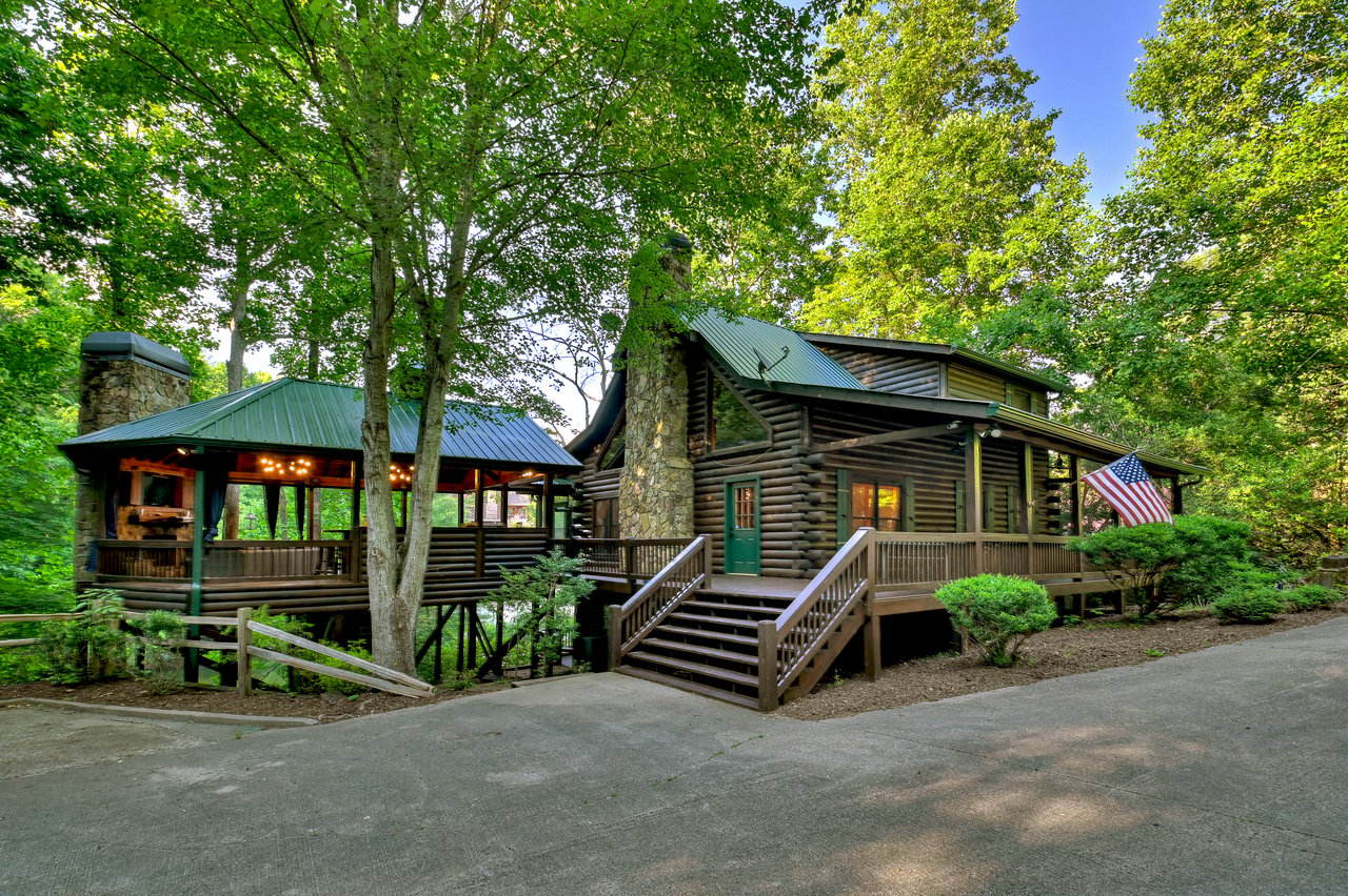 Log cabin home with stone chimney, covered porch, and American flag among leafy trees.