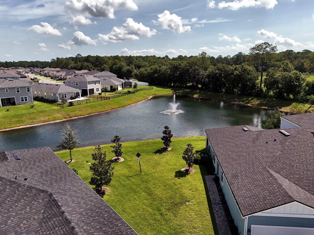 Aerial view of homes around a pond with a fountain and wooded shoreline.