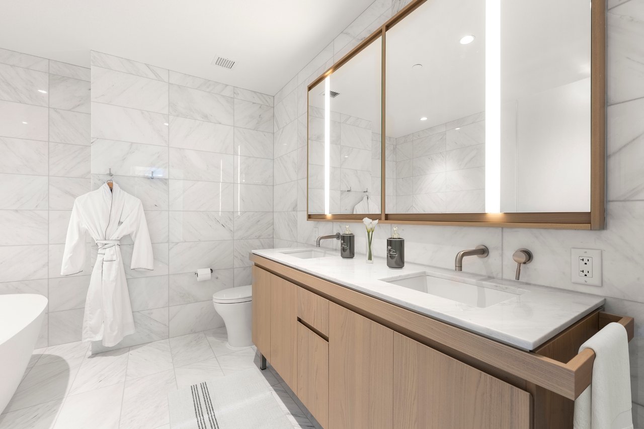 Modern marble bathroom with a double-sink wood vanity and a white robe hanging on the wall.