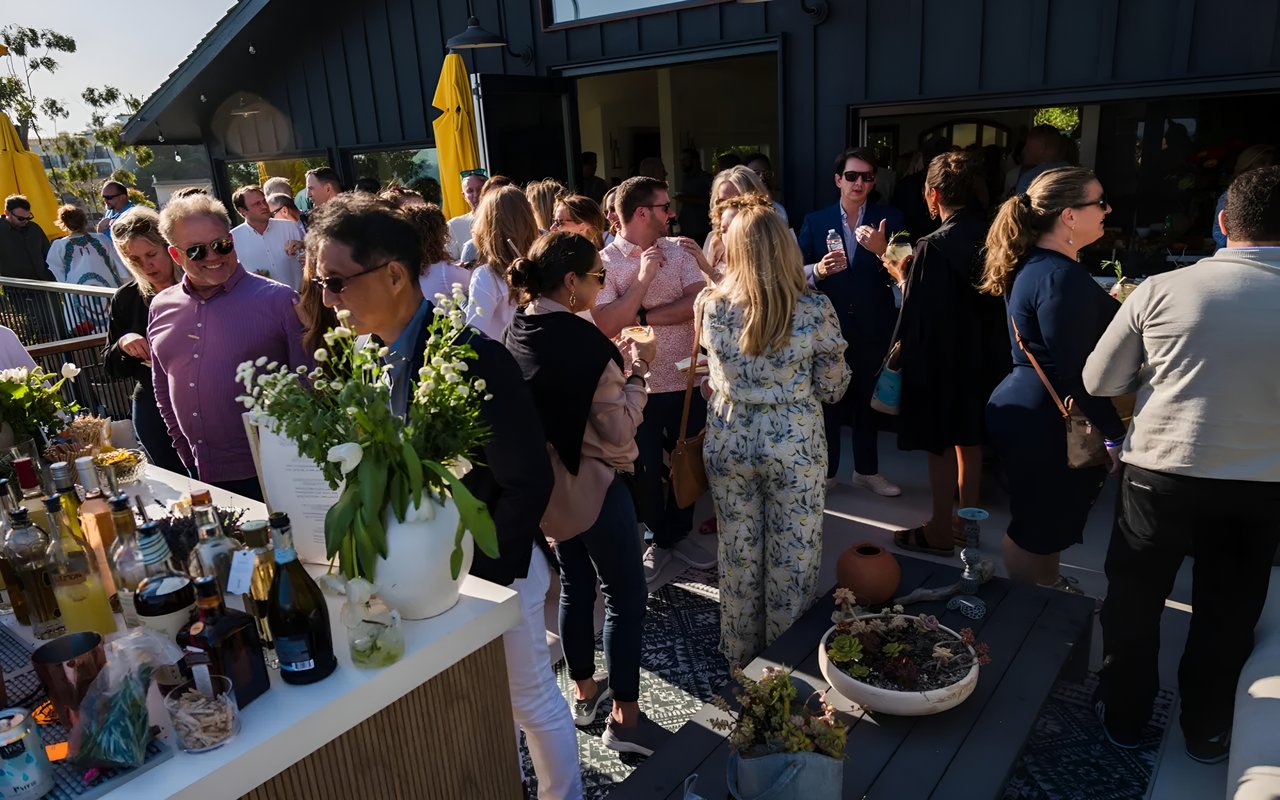  A crowded outdoor gathering with people socializing, with a bar and tables on a deck. The sun shines brightly.