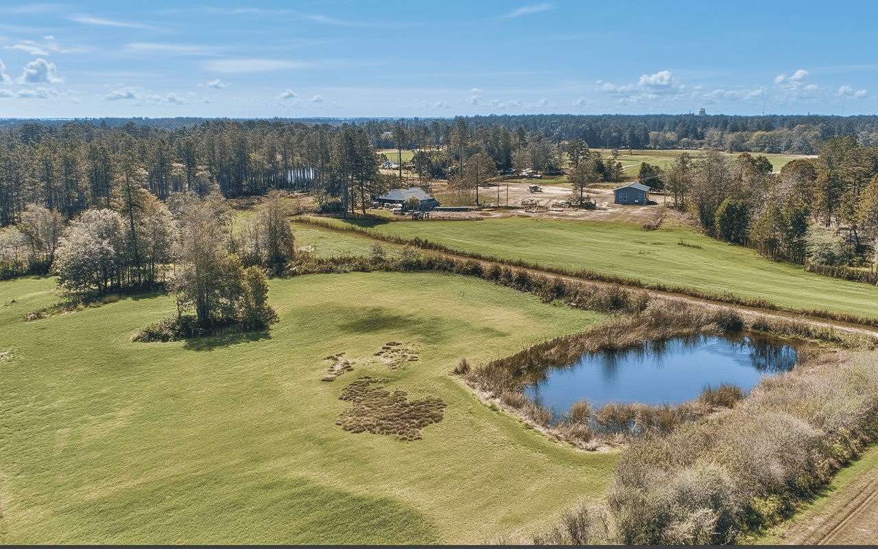 Aerial view of grassy acreage with a pond, wooded edges, and scattered buildings.